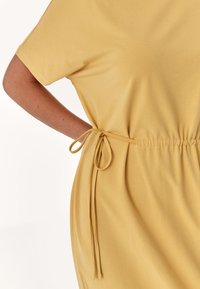 Gelbes, kurzärmliges Kleid mit elastischem Bund und gebundenem Detail. Weicher Stoff mit glatter Textur und lässiger Passform.