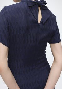 Femme portant une robe texturée bleu foncé à manches courtes avec un nœud noué au niveau de l'encolure arrière, dévoilant une petite ouverture en forme de trou de serrure.