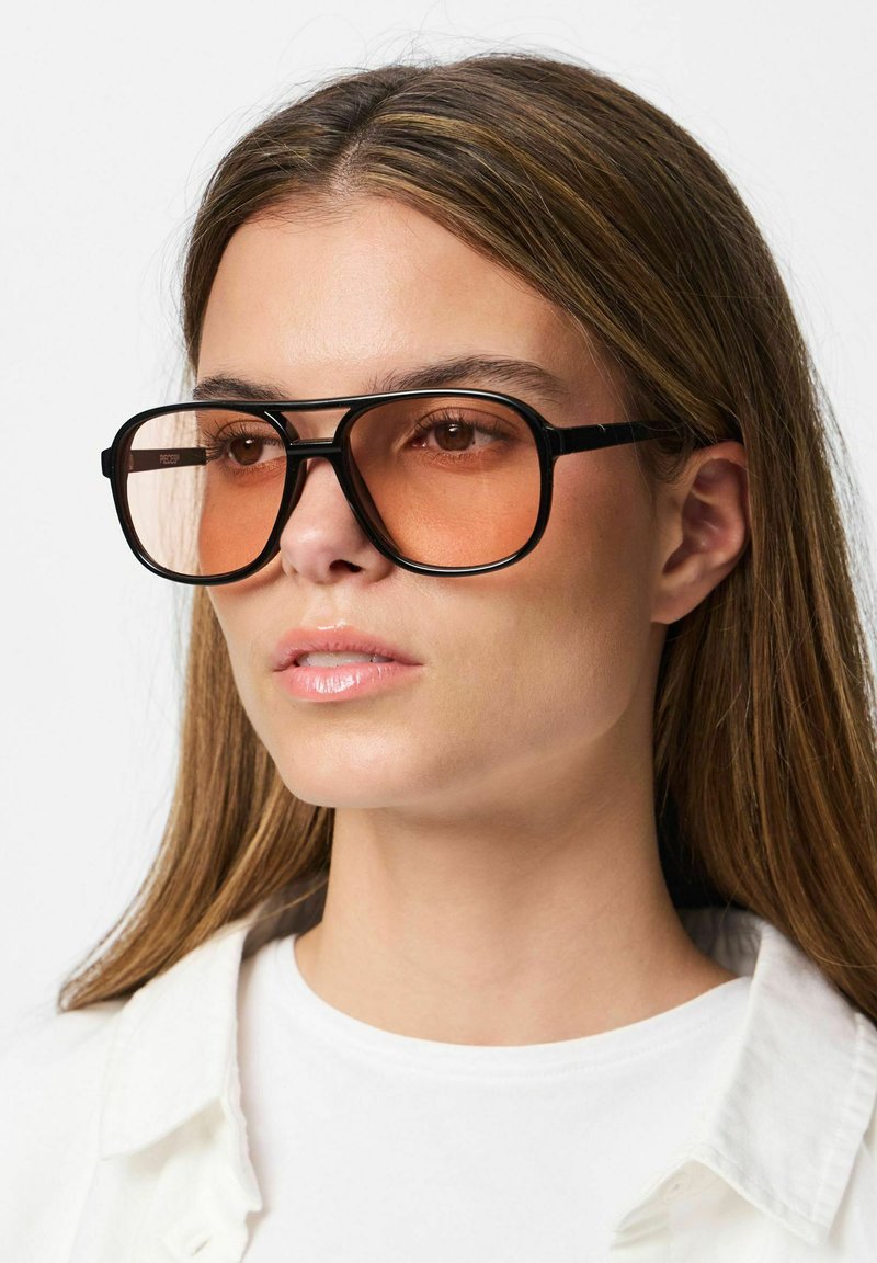 Femme aux longs cheveux bruns portant des lunettes de soleil surdimensionnées à monture noire avec des verres teintés orange et une chemise blanche.