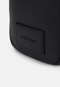 Geantă sintetică neagră cu o textură netedă și o patch din piele rectangulară cu un logo embosat. Design simplu, elegant.