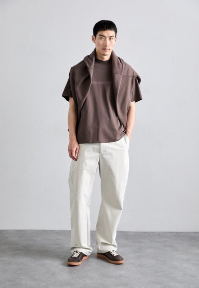 Braunes übergroßes T-Shirt mit einem horizontalen Streifenmuster, kombiniert mit cremefarbenen, plissierten Hosen und braunen Sneakers mit Wildlederoptik.