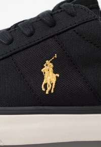 Svart canvas-sneaker med en guldfärgad broderad polo-spelarlogotyp, texturerat tyg och en vit gummisula för kontrast.