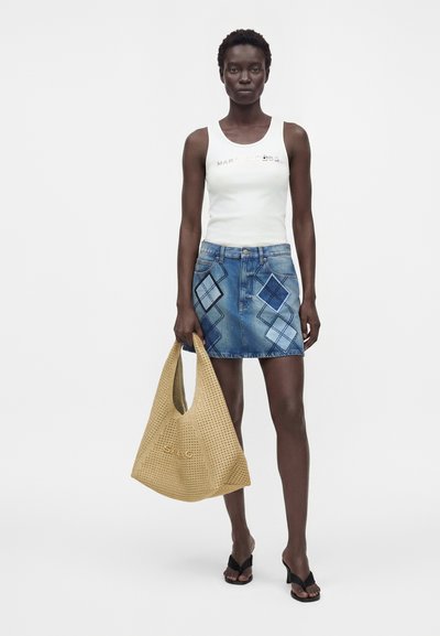 Femme portant un top blanc sans manches et une jupe en jean bleue avec des détails de patch, tenant un grand sac beige tissé et portant des sandales noires.