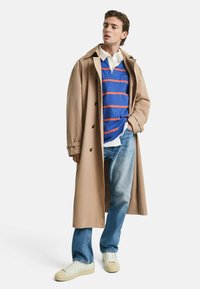 Beige trenchcoat, blå stripete genser med oransje striper, hvit skjorte, lyseblå jeans, kremfargede joggesko. Glatt stoff, avslappet passform.
