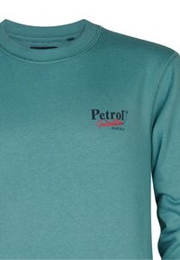 Sudadera de color verde azulado hecha de tela suave, con cuello de barco y puños acanalados. En la parte frontal tiene un logo en texto negro y rojo.