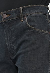 Tmavé džíny s vysokým pásom, kontrastným šitím, kovovými detailmi a klasickým päť-pocket štýlom.