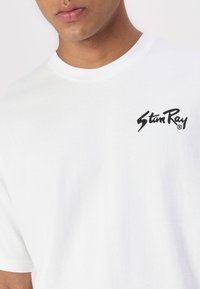 White cotton t-shirt featuring a black logo on the left chest area. The fabric appears smooth with a classic crew neckline and short sleeves.