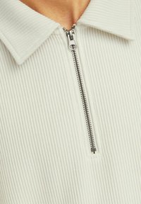 Top blanco acanalado con cremallera frontal y cuello tipo solapa. Textura suave con líneas verticales.