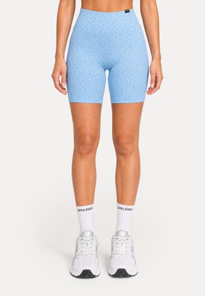 Femme portant un short de cycliste taille haute bleu clair à motifs, des chaussettes blanches avec texte et des chaussures de running blanches, debout devant un fond uni.