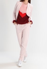 Femme portant un costume rose pâle avec un pull à chevrons rouge et bordeaux et des baskets blanches, les mains dans les poches, se tenant devant un fond clair.