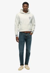 Jasnoszara bluza z kapturem z kieszenią z przodu i granatowym napisem; niebieskie dżinsy; kremowe sneakersy; dopasowany fason; ściągacze z ribbingu w granatowe paski.