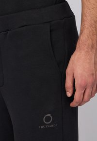 Czarne dresy z miękkiego, teksturowanego materiału; mają boczne kieszenie i subtelne, srebrne logo Trussardi blisko pasa.