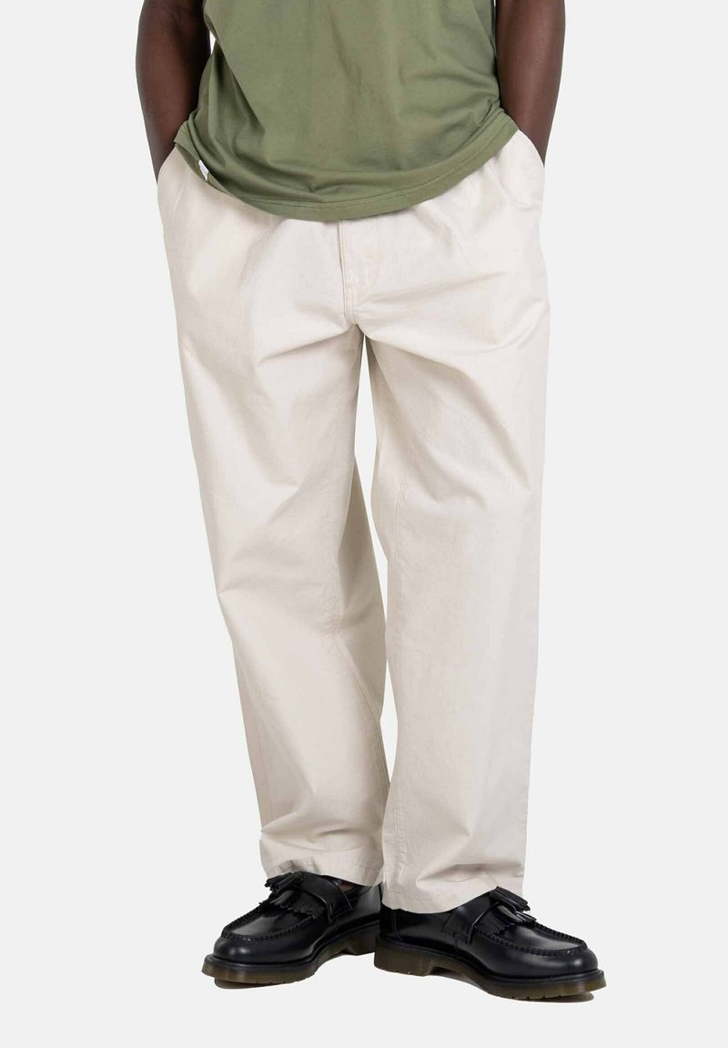 Light beige cotton pants with a relaxed fit, featuring side pockets, paired with black leather shoes and an olive green shirt.