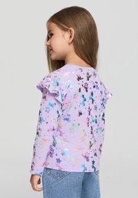 Fioletowa koszula z długim rękawem z iryzującymi wzorami jednorożców i gwiazdek, z falbankami na ramionach oraz okrągłym dekoltem.