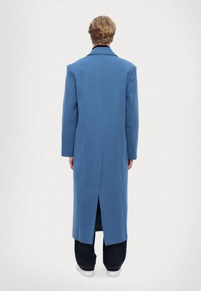 Dlhý, štruktúrovaný modrý kabát z vlny, s jedným rozventením na chrbte a klopom, nosený cez tmavé nohavice a biele topánky.