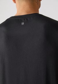 Juoda marškinėliai su apvalia apykakle ir trumpomis rankovėmis, pagaminti iš lygaus audinio. Nugaros viršuje, netoli apykaklės, yra mažas baltas logotipas.