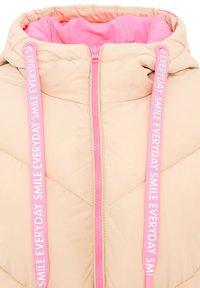Beige Pufferjacke mit pinkem Innenfutter und Reißverschluss. Mit rosa Zugbändern und dem Text "SMILE EVERYDAY". Diamantgestepptes Muster auf der gesamten Fläche.