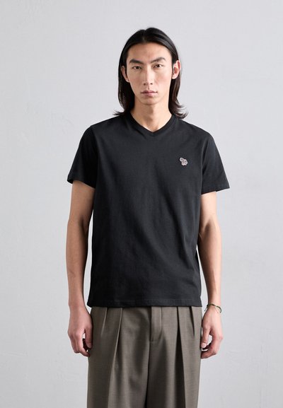 Tricou negru din bumbac cu decolteu în V, având un logo mic, colorat pe pieptul stâng. Modelul poartă pantaloni largi, cu pliuri, de culoare maronie deschisă.