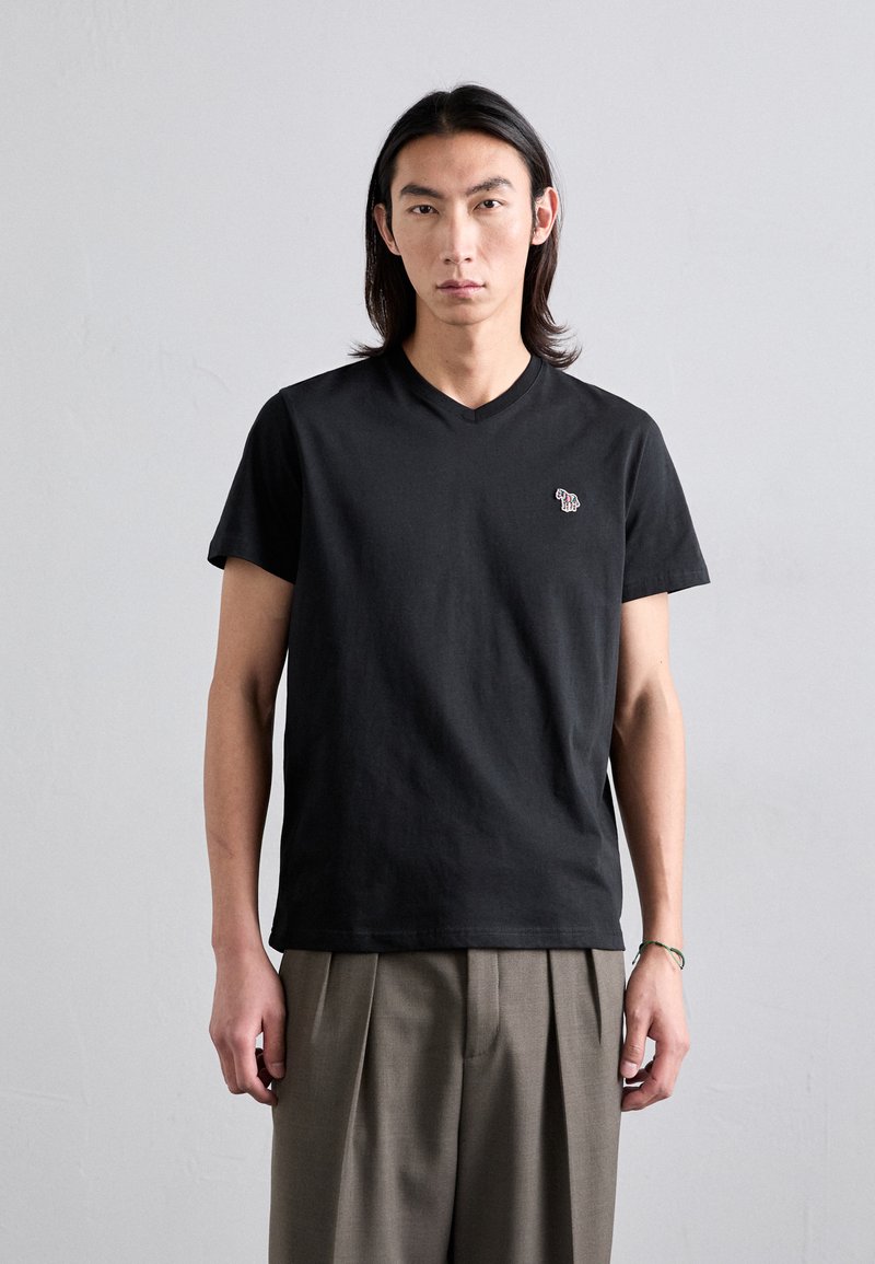 T-shirt V en coton noir avec un petit logo coloré sur la poitrine gauche. Le modèle porte un pantalon plissé léger de couleur marron clair.
