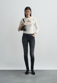 White knitted sweater with a collar detail, dark denim skinny jeans, black handbag, and black pointed shoes against a plain background.