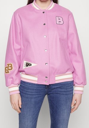 Osoba w jasnoróżowej kurtce varsity z białymi paskami na mankietach i dole, z naszywkami w formie liter oraz w niebieskich dżinsach, stojąca na tle jednolitego tła.