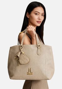 Borsa tote in pelle beige con due manici allungati, arricchita da un piccolo charm a forma di cuore e accessori in metallo color oro. Logo in rilievo sulla parte anteriore.