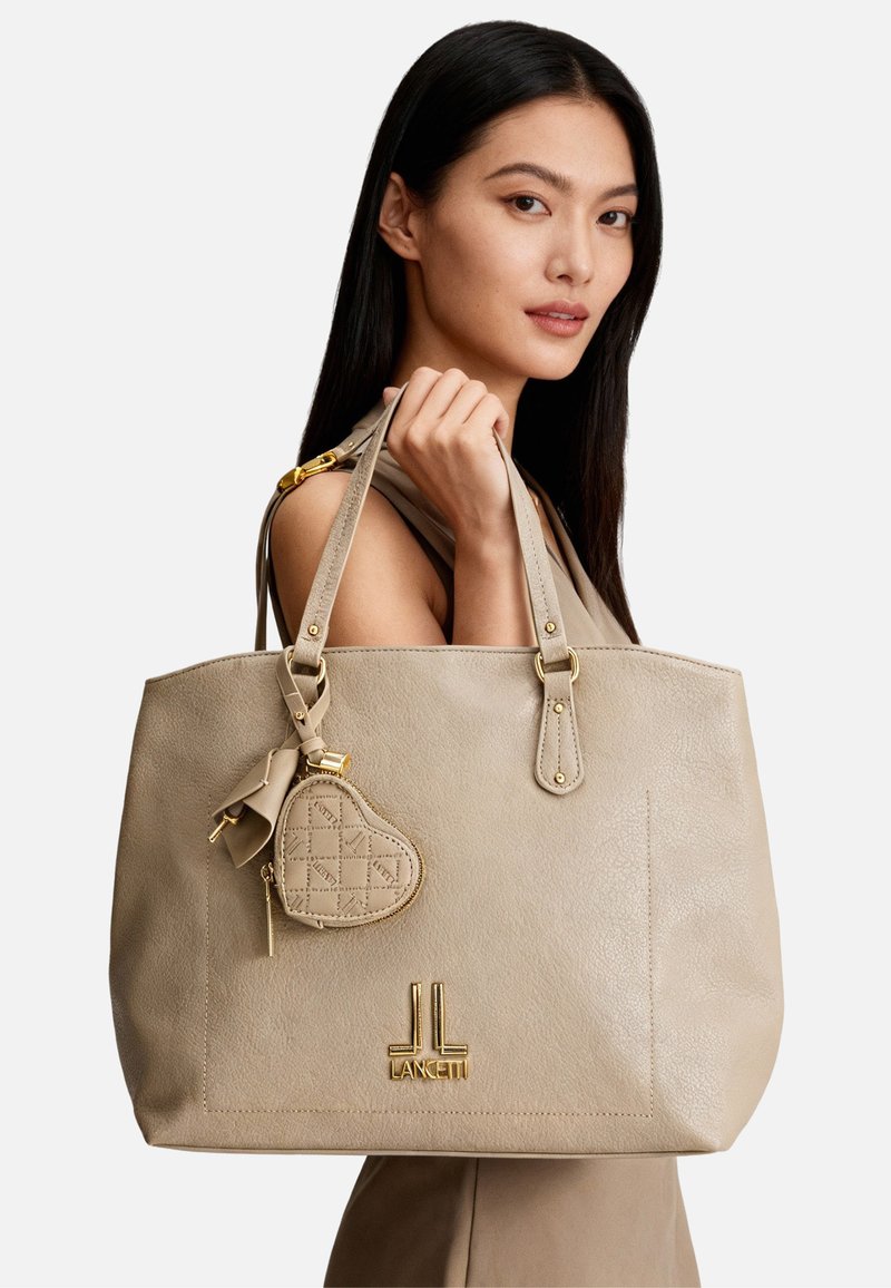 Borsa tote in pelle beige con due manici allungati, arricchita da un piccolo charm a forma di cuore e accessori in metallo color oro. Logo in rilievo sulla parte anteriore.