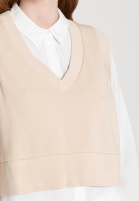 Chaleco sin mangas de punto beige con escote en V y dobladillo acanalado, superpuesto a una camisa blanca con cuello. Textura suave, corte moderno.