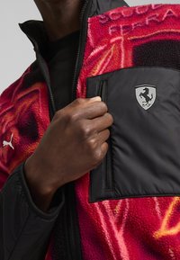 Fleece jacket with a vibrant red and pink pattern, featuring a black chest pocket and silver Ferrari logo on the front.