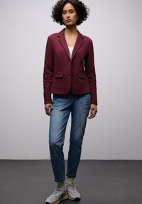 Blazer en bordeaux texturé avec deux poches avant, assorti à un jean bleu clair et des baskets grises. Col simple et détail de fermeture à boutons.