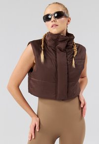 Smeđi skraćeni puffer prsluk s visokim ovratnikom, bez rukava i prednjim zatvaračem. Materijal je gladak s blagim sjajem.