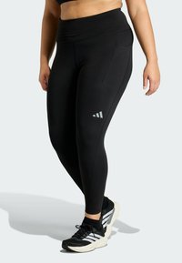 Black leggings with a high waistband, featuring a side pocket and a reflective logo detail on the left leg. Worn with black athletic shoes.