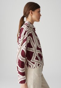 Camicia a maniche lunghe di colore bordeaux e bianco sporco, con colletto e design corto, caratterizzata da forme geometriche astratte e linee.