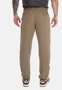 Pantaloni leggeri color khaki realizzati in tessuto con finitura testurizzata. Presentano due tasche posteriori con bottoni. Indossati con sneakers marroni e bianche.