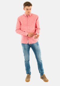 Chemise à manches longues boutonnée en tissu rose clair, dotée d'un col classique et d'un ourlet droit, portée avec un jean bleu clair et des baskets brunes.