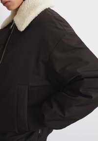 Svart bomberjacka med en fluffig krämfärgad krage, ribbade ärmslut och sidofickor, som har en slät yta och en avslappnad passform.