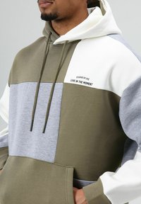 Olivengroene, grijze en witte hoodie met kleurvlakken, voorzien van een kangoeroezak, trekkoord in de capuchon en bedrukte tekst op de borst.