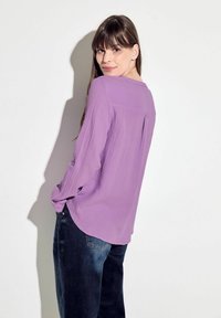 Blusa larga de manga larga en color púrpura con tejido texturizado, escote redondo y dobladillo circular. Combinada con unos jeans de mezclilla azul oscuro.