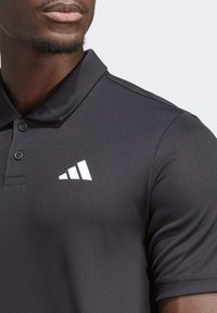 Schwarzes Polohemd aus glattem Stoff mit Kragen, ausgestattet mit drei Knöpfen und einem auffälligen weißen Markenlogo auf der linken Brust.