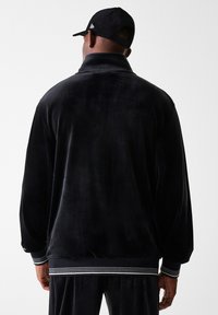 Veste en velours noir avec col montant, poignets et ceinture côtelés ornés de bandes blanches. Texture lisse et coupe décontractée.