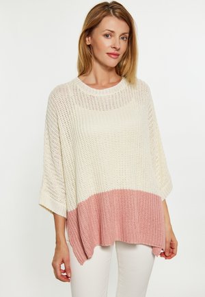 Kvinde iført en løs strikket sweater i creme med et bredt lyserødt bånd i bunden, stående mod en ensfarvet lys baggrund.