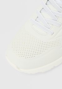 Adidaș alb cu partea superioară sintetică perforată, șireturi plate și guler căptușit. Prezintă un vârf rotunjit și o talpă texturată pentru aderență.