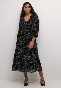 Vestido negro con lunares hecho de material ligero, con escote en V, mangas largas abullonadas y una falda fluida hasta los tobillos.