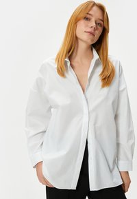 Camicia bianca con bottone, dal fit morbido, maniche lunghe e colletto classico. Tessuto liscio, con bottoni frontali e spacchi laterali.