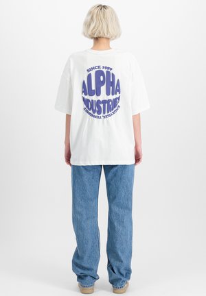 Bílý oversize tričko s velkým fialovým potiskem "ALPHA INDUSTRIES" na zadní straně, v kombinaci se světle modrými denimovými kalhotami.