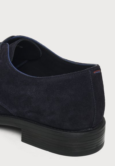 Gros plan sur le talon d'une chaussure habillée en daim bleu marine avec une semelle noire superposée et un subtil détail de couture rouge et bleue à l'arrière.