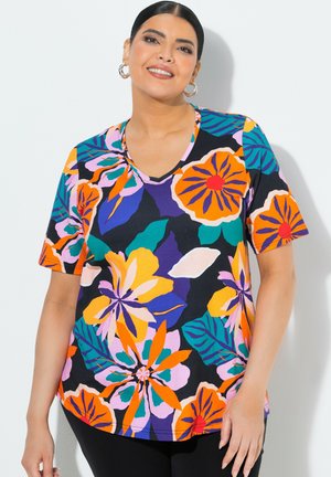Femme souriante aux cheveux foncés portant une chemise à manches courtes et à motifs floraux vibrants orange, violet, rose et vert, avec des boucles d'oreilles créoles.