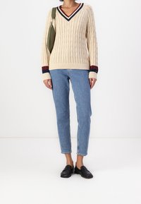 Beige kaapeknitti neuletakki V-pääntiellä, jossa tummansinisiä ja punaisia raidallisia yksityiskohtia hihoissa. Yhdistettynä vaaleansiniset istuvat farkut ja mustat kengät.