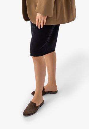 Kobieta ubrana w płaszcz koloru wielbłądziego, czarną spódnicę za kolano i brązowe wsuwane buty, stojąca z jedną ręką spoczywającą na udzie.