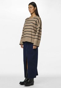 Pull en grosse maille beige à rayures, coupe ample avec un ourlet à côtes, associé à une longue jupe en denim foncé avec une fente à l'avant, complété par des bottes à talons noires.